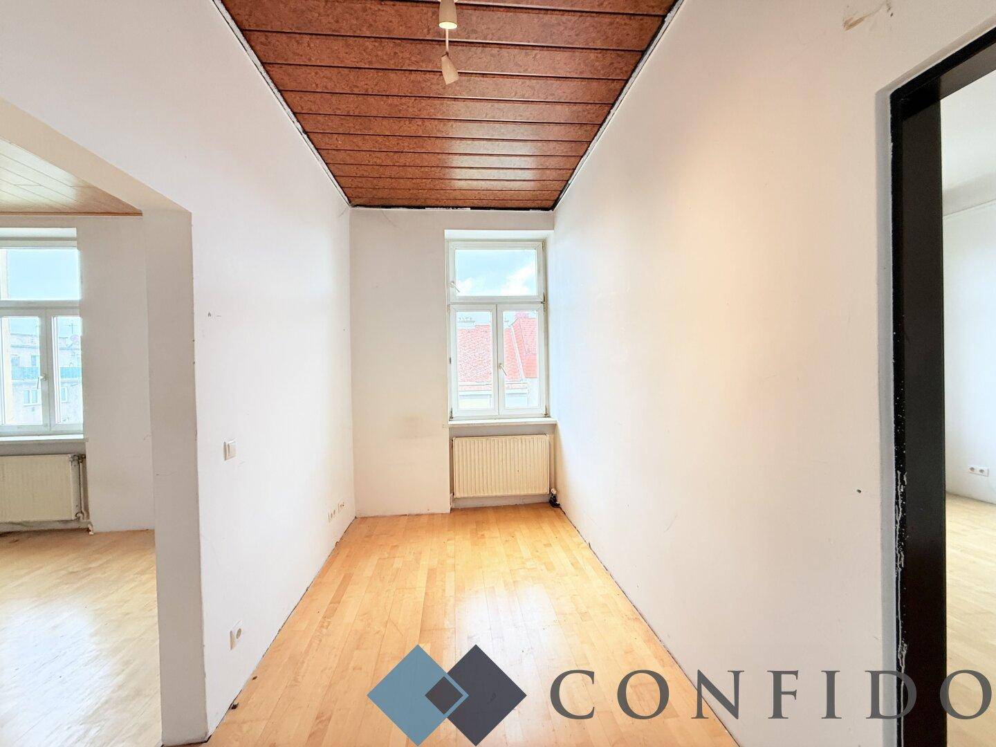 Schmaler Raum mit Holzdeckenverkleidung, Parkettboden und Fenster, verbindet andere Bereiche.