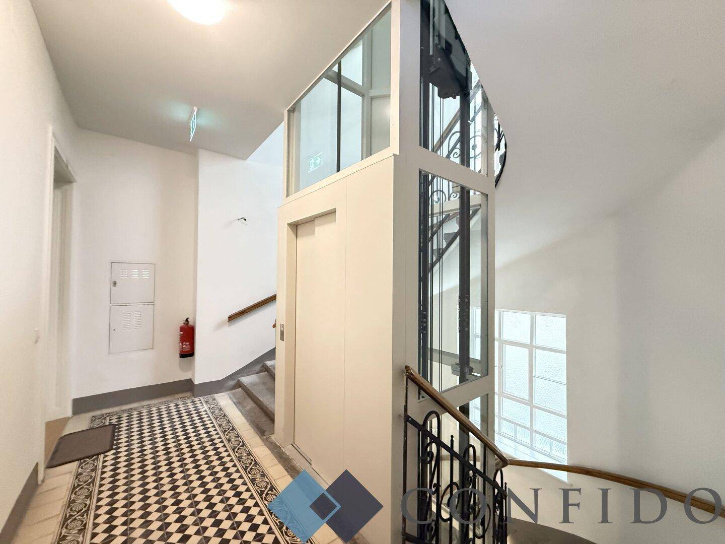 Historisches Treppenhaus mit modernem Glasaufzug und Schachbrettmuster-Fliesenboden.
