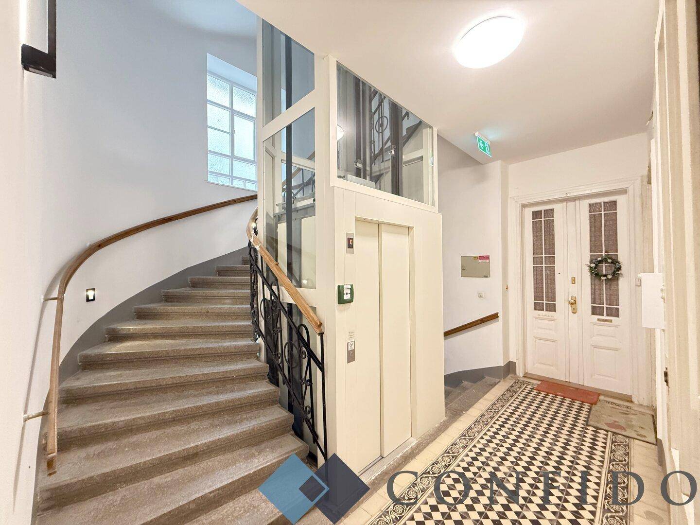 Elegantes Treppenhaus mit geschwungener Treppe, modernem Aufzug und Fliesenboden.