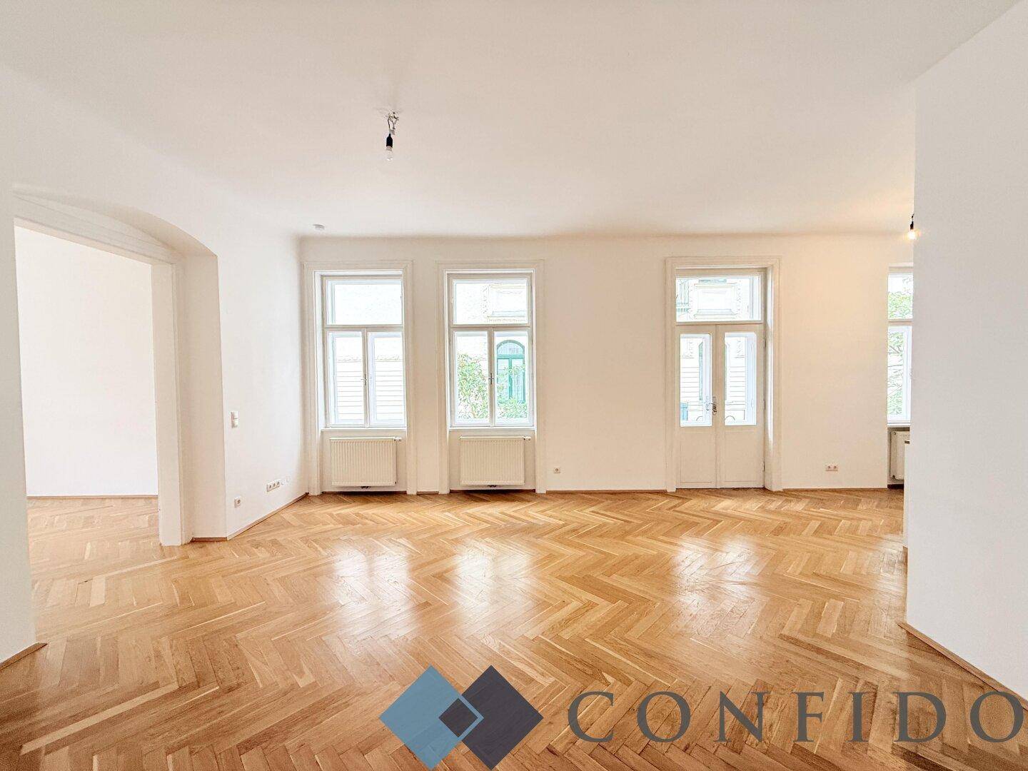Großes, helles Zimmer mit Fischgrätparkett und hohen Fenstern, ideal als Wohnzimmer.