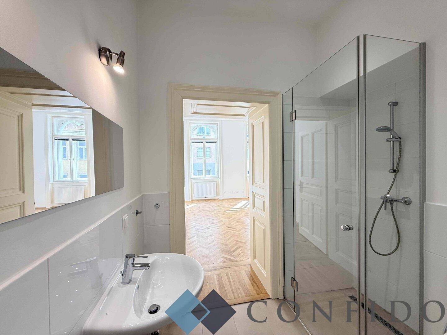 Badezimmer mit Duschkabine, Einzelwaschbecken und Blick in ein angrenzendes Zimmer.