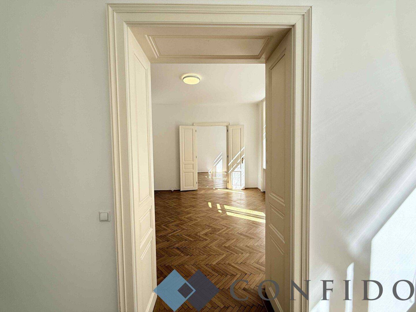 Blick durch eine hohe Flügeltür in ein helles Zimmer mit Fischgrätparkett und Tageslicht.
