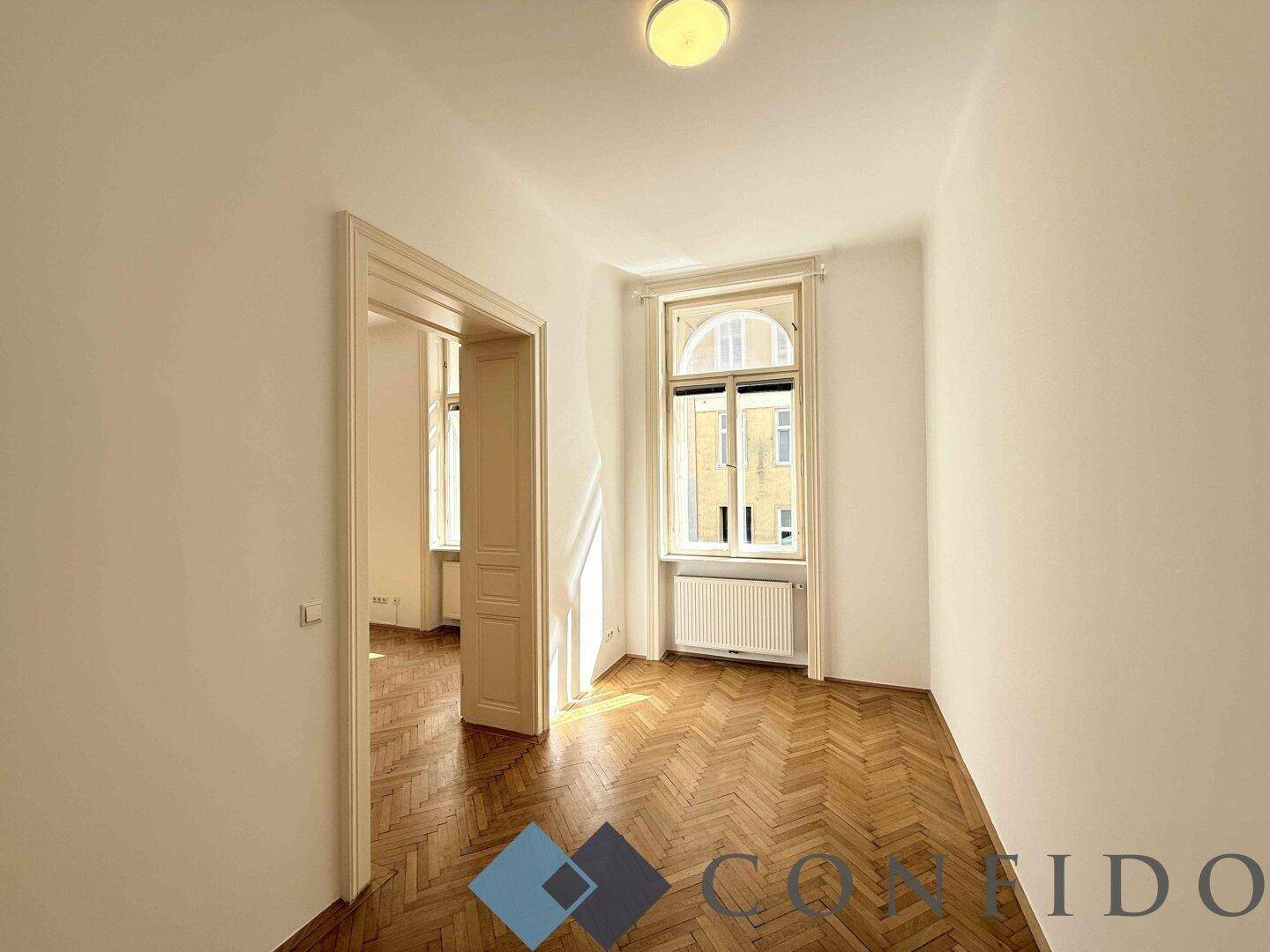 Helles Zimmer mit Fischgrätparkett und einem hohen Fenster, das viel Tageslicht bietet.
