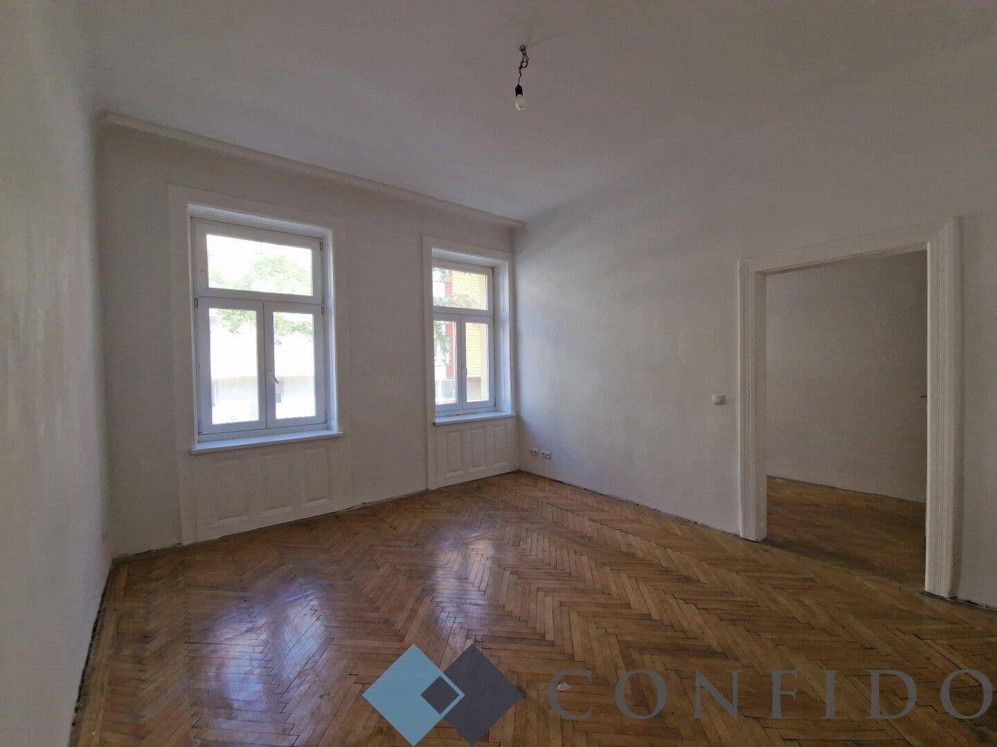 Helles Zimmer mit zwei großen Fenstern und klassischem Fischgrätparkett, ideal für Wohnzwecke.