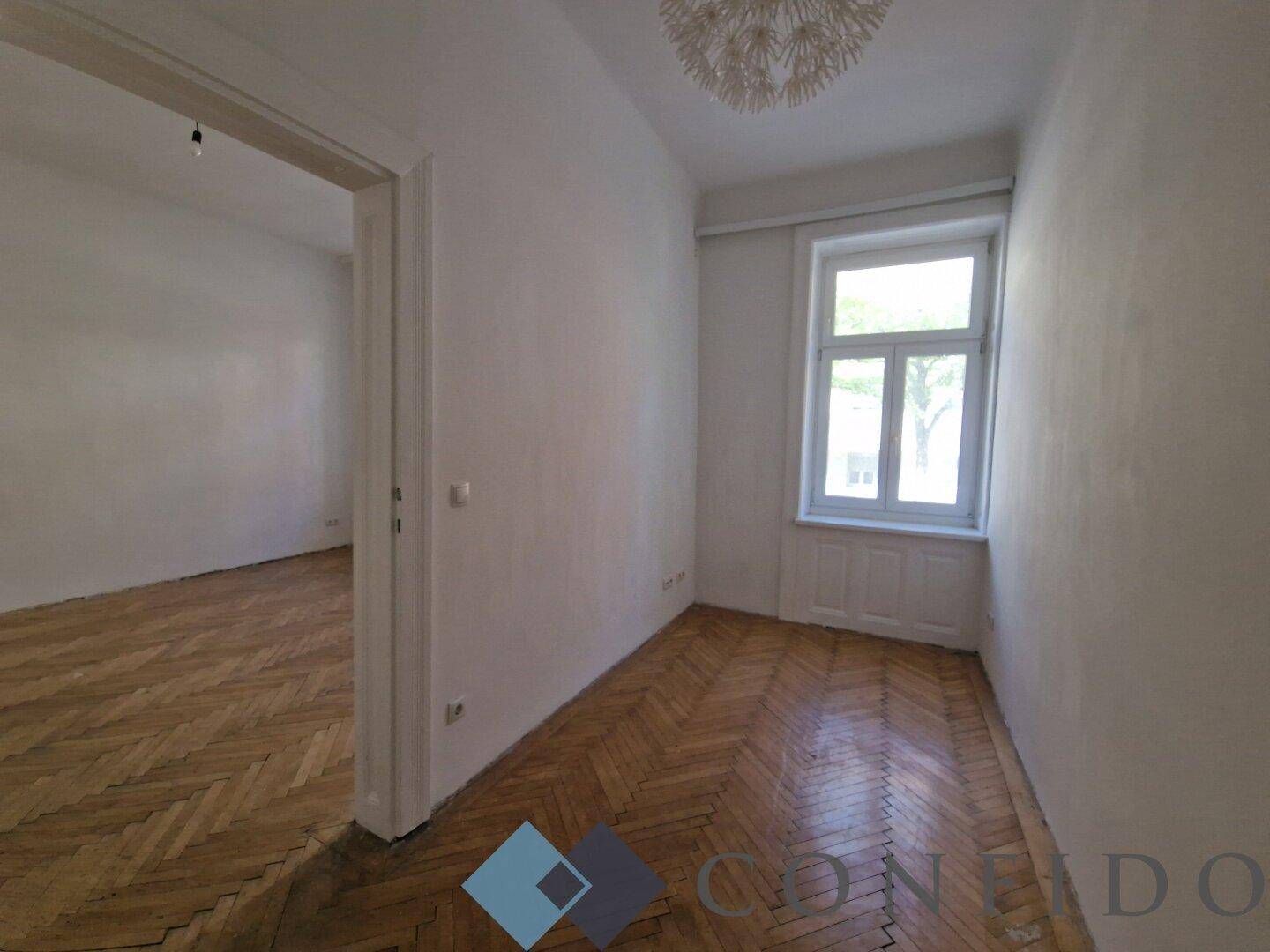 Schmaler Raum mit Fischgrätparkett und einem Fenster, das Tageslicht hereinlässt.