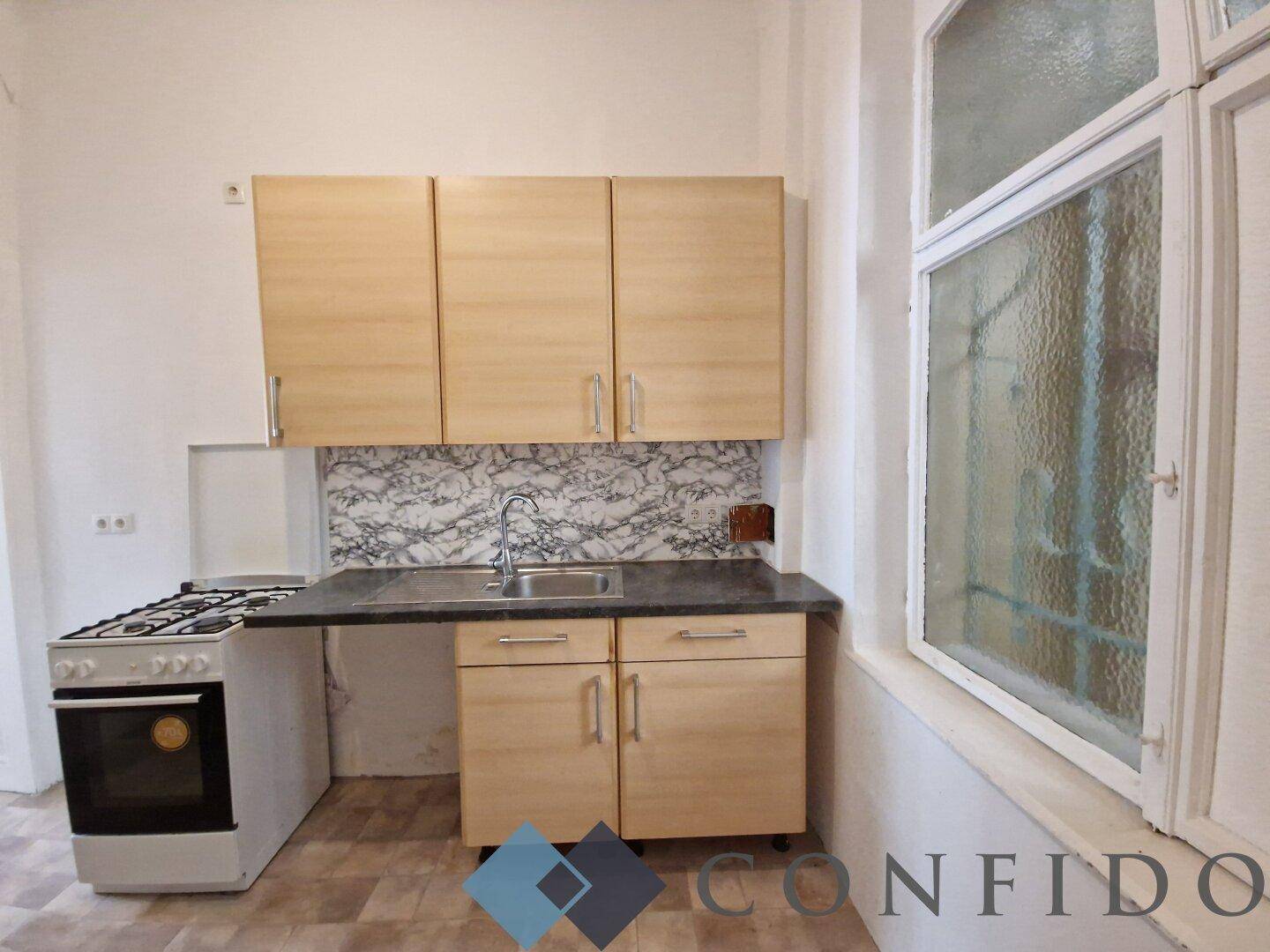 Einfache Küche mit Gasherd, Spüle und Holzschränken unter einem großen Fenster.