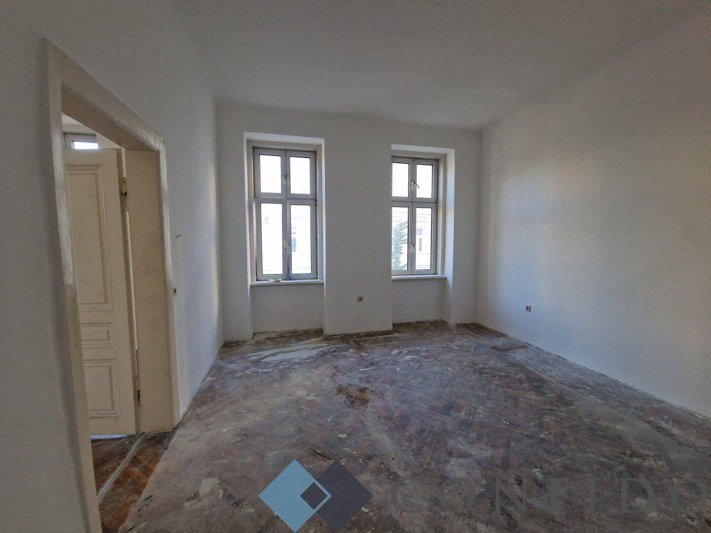 Geräumiger Wohnraum im Rohbau mit zwei hohen Fenstern, ideal für individuelle Gestaltung.