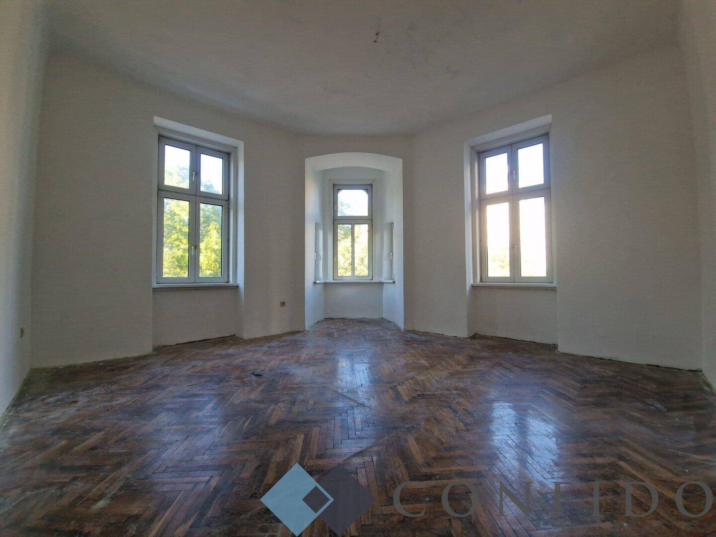 Heller Raum mit Erkerfenstern und altem Fischgrätparkett, bereit für eine Sanierung.