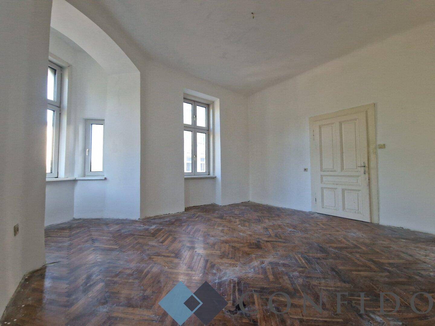 Wohnraum mit Erkerfenstern, abgenutztem Parkettboden und einer traditionellen Holztür.