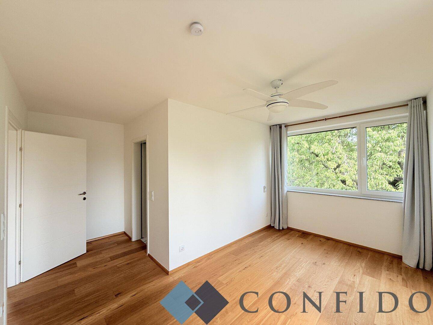 Helles Schlafzimmer mit Holzboden, Deckenventilator und großem Fenster mit Blick ins Grüne.