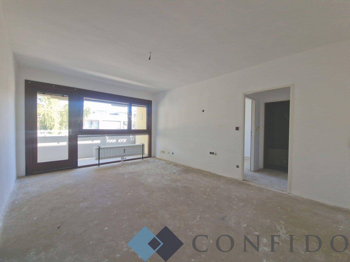 Unmöblierter Wohnbereich mit großen Fenstern und Zugang zu einem Balkon, bereit für die Renovierung.
