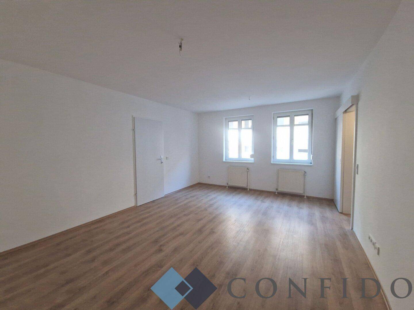 Leeres Wohnzimmer mit hellem Laminatboden und zwei Fenstern, bereit zur individuellen Gestaltung.
