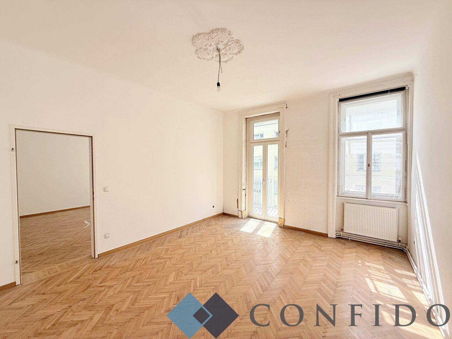 Helles, leeres Zimmer mit Fischgrätparkett, Stuckdecke und Zugang zu einem französischen Balkon.