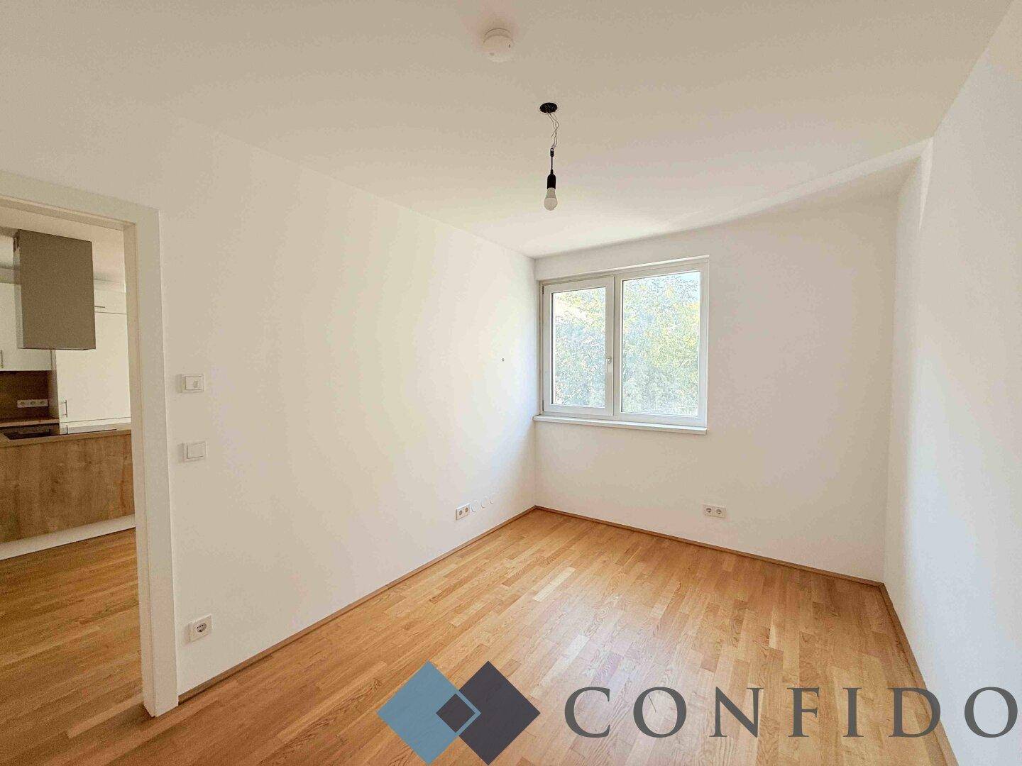 Helles Schlafzimmer mit Parkettboden und einem Fenster mit Blick ins Grüne.
