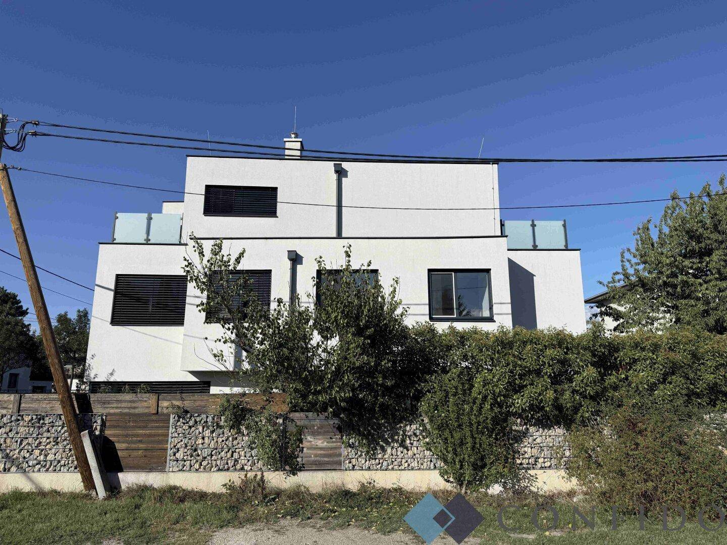 Moderne weiße Fassade des Gebäudes mit dunklen Fenstern und Gabionenzaun im Vordergrund.