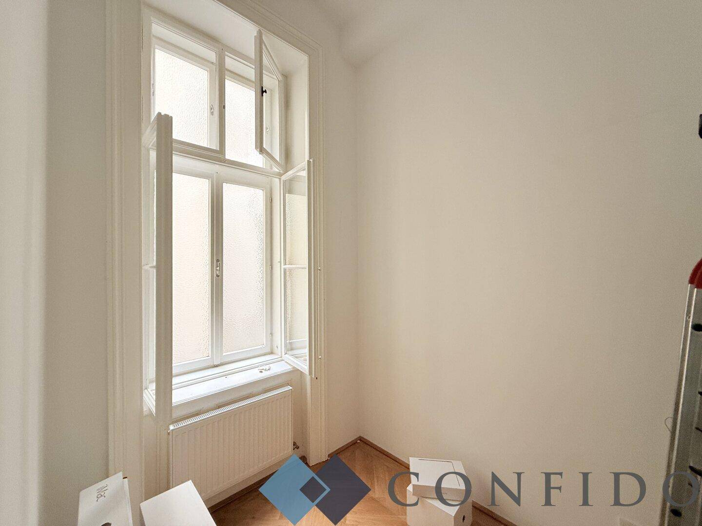 Kleiner Raum mit einem hohen Fenster, das viel Tageslicht hereinlässt, und Holzboden.