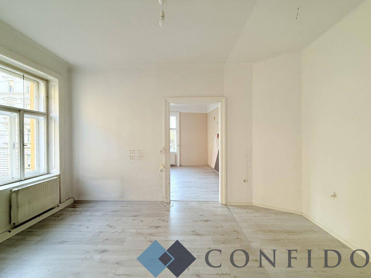 Helles Zimmer mit Laminatboden und einem Fenster, das einen Blick in einen angrenzenden Raum ermöglicht.