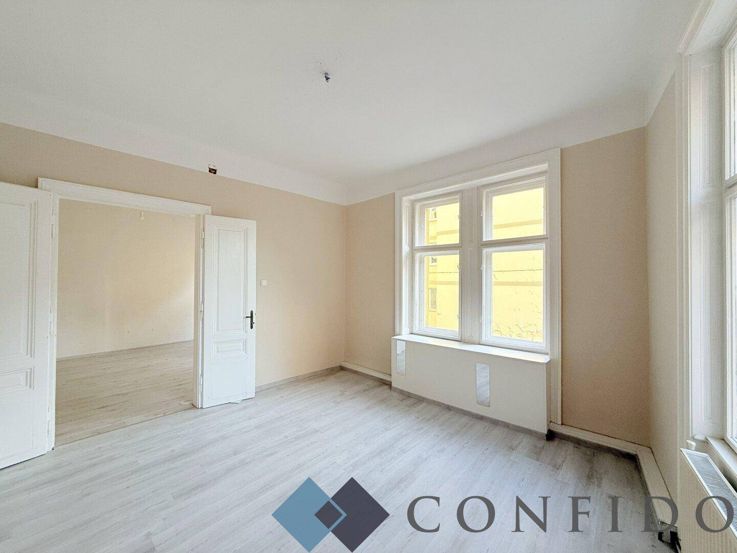 Helles Zimmer mit beigen Wänden, Laminatboden und einem großen Fenster, das viel Tageslicht hereinlässt.