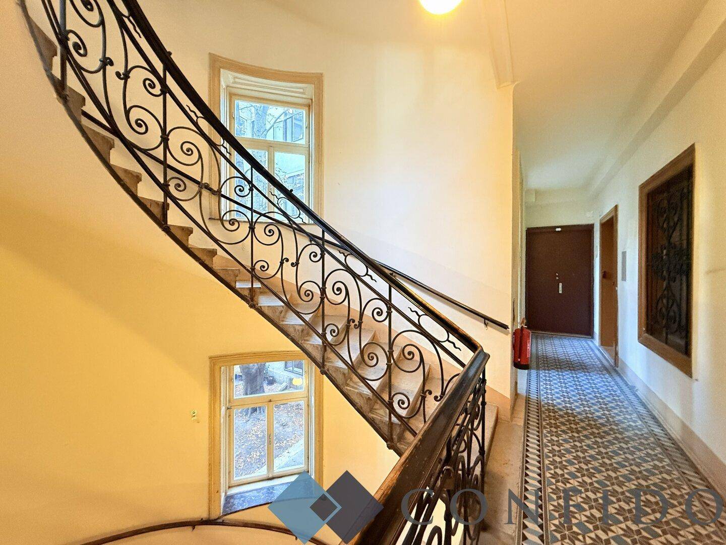 Historisches Treppenhaus mit schmiedeeisernem Geländer und gemustertem Fliesenboden, hell durch Fenster.