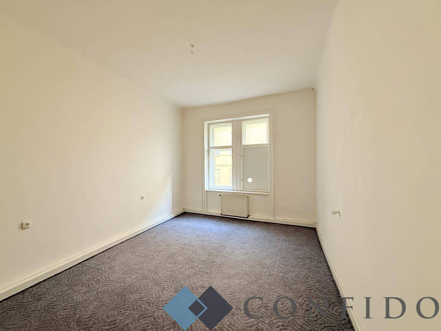 Gemütliches, leeres Zimmer mit grauem Teppichboden und einem Fenster, das für natürliches Licht sorgt.