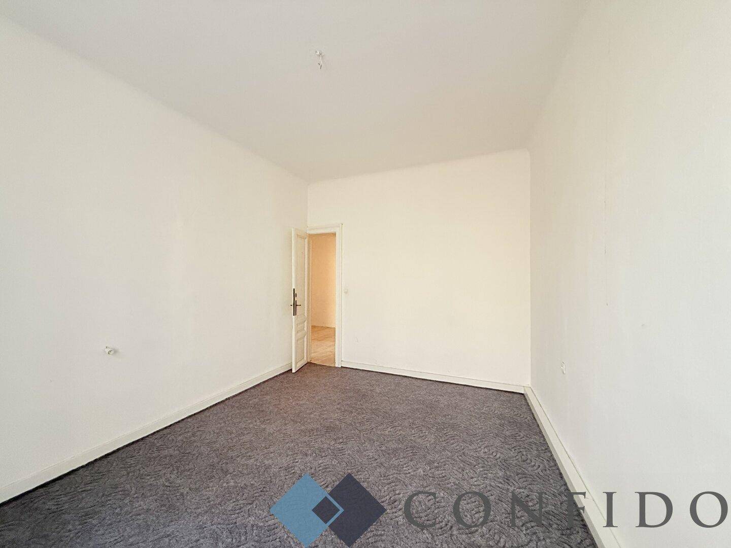 Leeres Zimmer mit grauem Teppichboden und weiß gestrichenen Wänden, das durch eine offene Tür zugänglich ist.