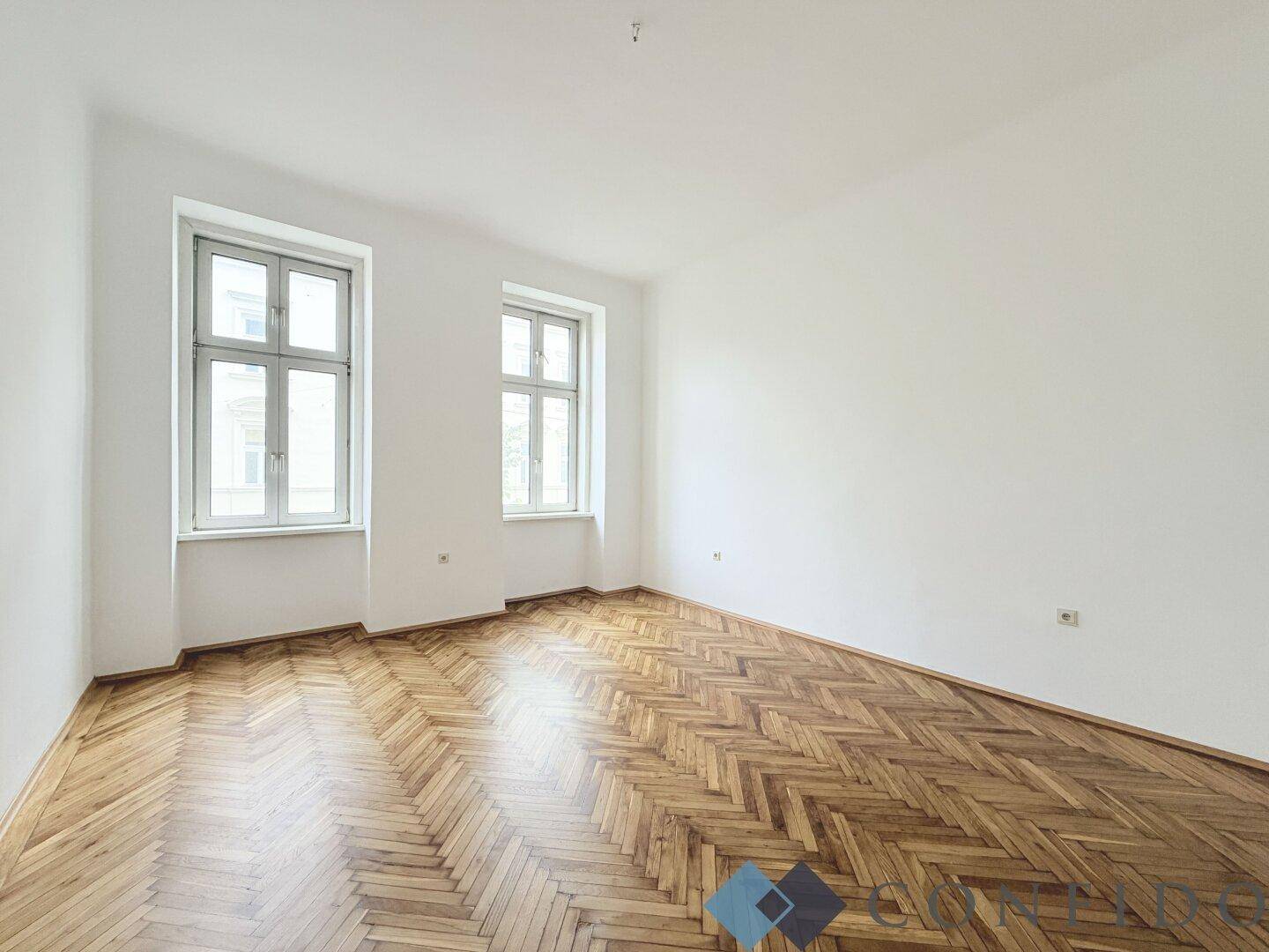 Helles Zimmer mit Fischgrätparkett und zwei hohen Fenstern, bereit zur individuellen Gestaltung.