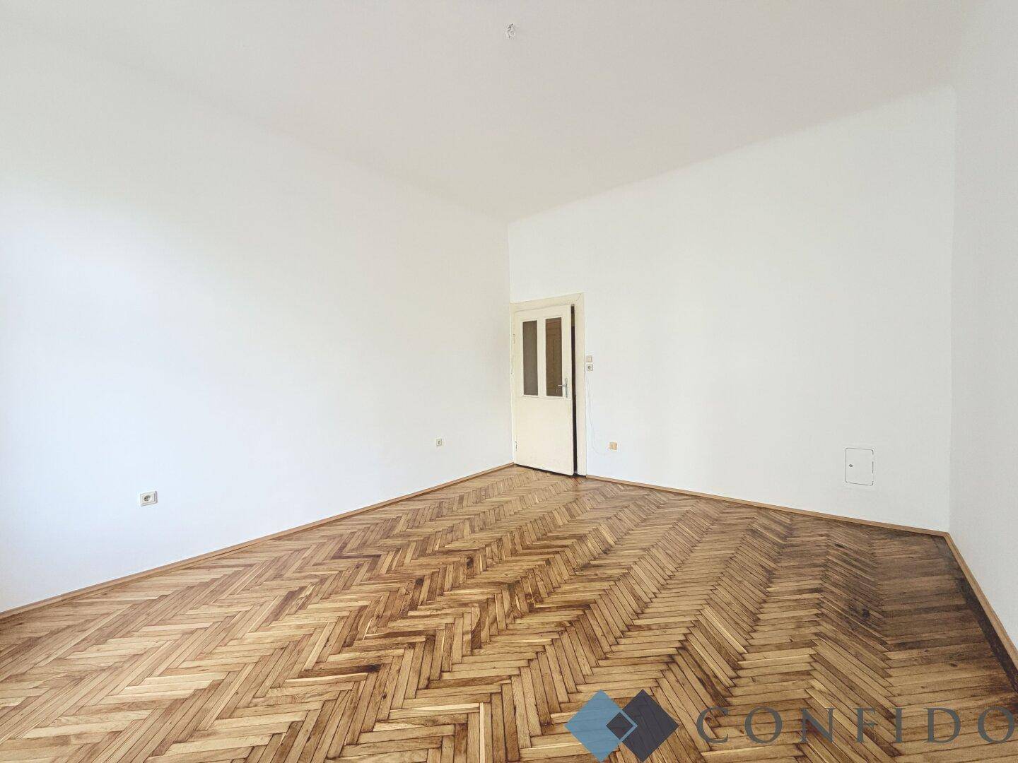 Leeres Zimmer mit Fischgrätparkett und einer Tür, bietet vielseitige Nutzungsmöglichkeiten.