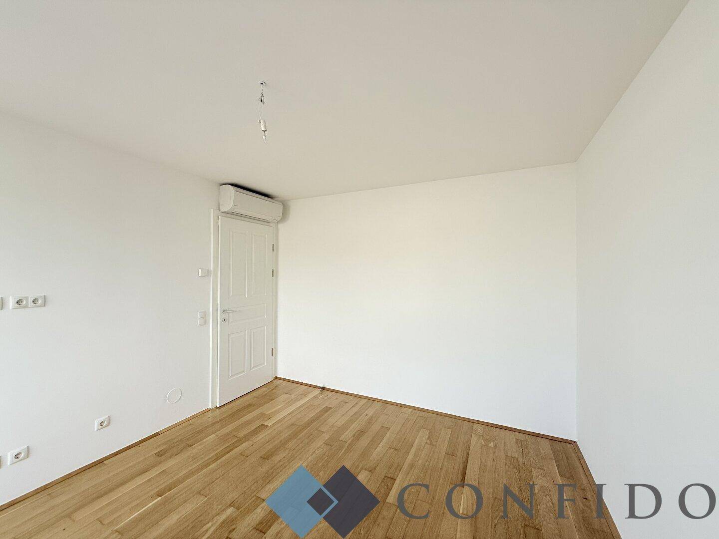 Leerer Raum mit Holzboden, weißer Tür und einer Klimaanlage an der Wand.