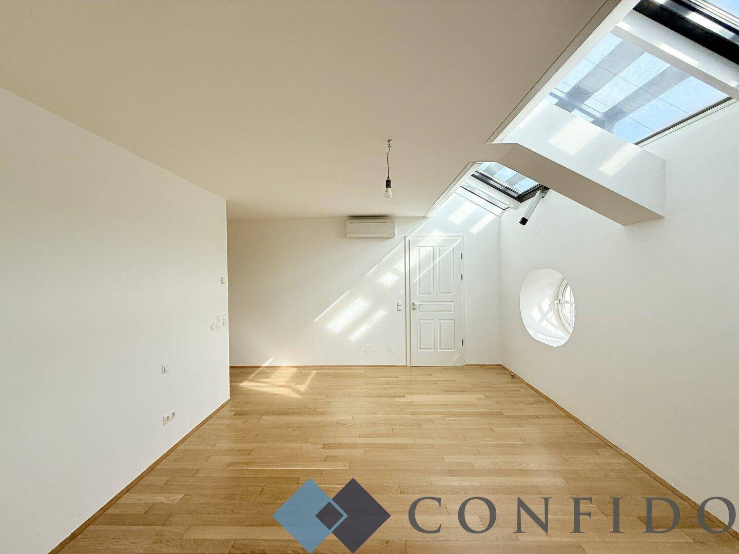 Helles Zimmer im Dachgeschoss mit Parkettboden, Dachfenstern und runden Fenstern.