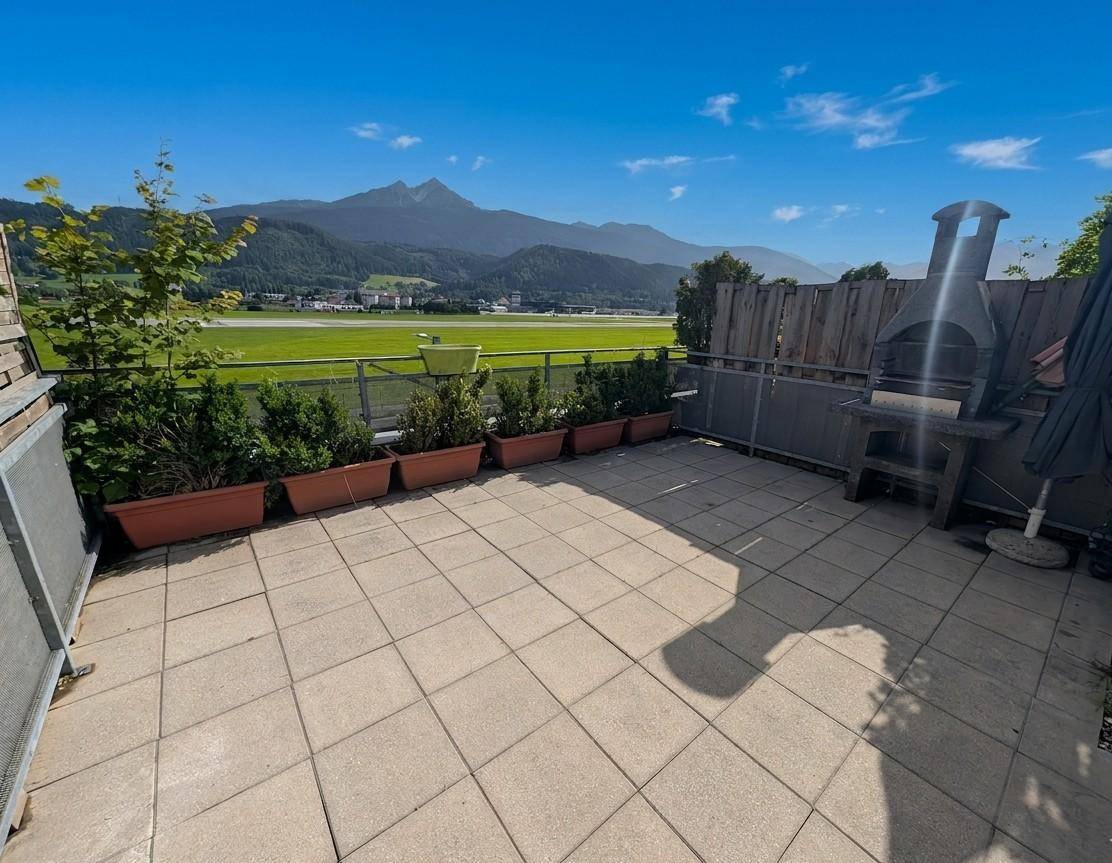 Geräumige Terrasse mit Bergblick, Grillplatz und Pflanzkübeln unter blauem Himmel.