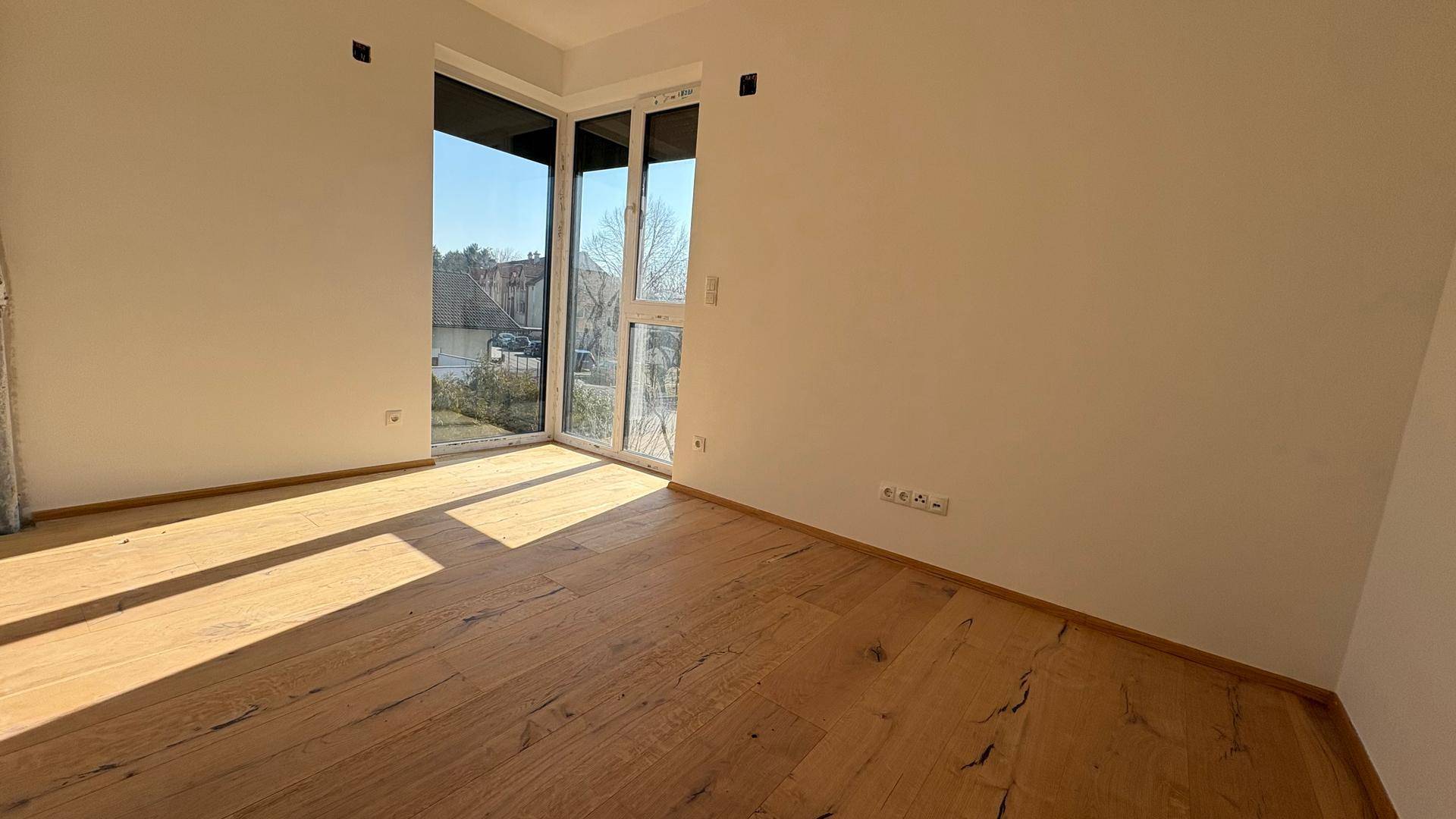 Helles Zimmer mit Holzboden und großen Fenstern, bietet viel Tageslicht und flexible Nutzungsmöglichkeiten.