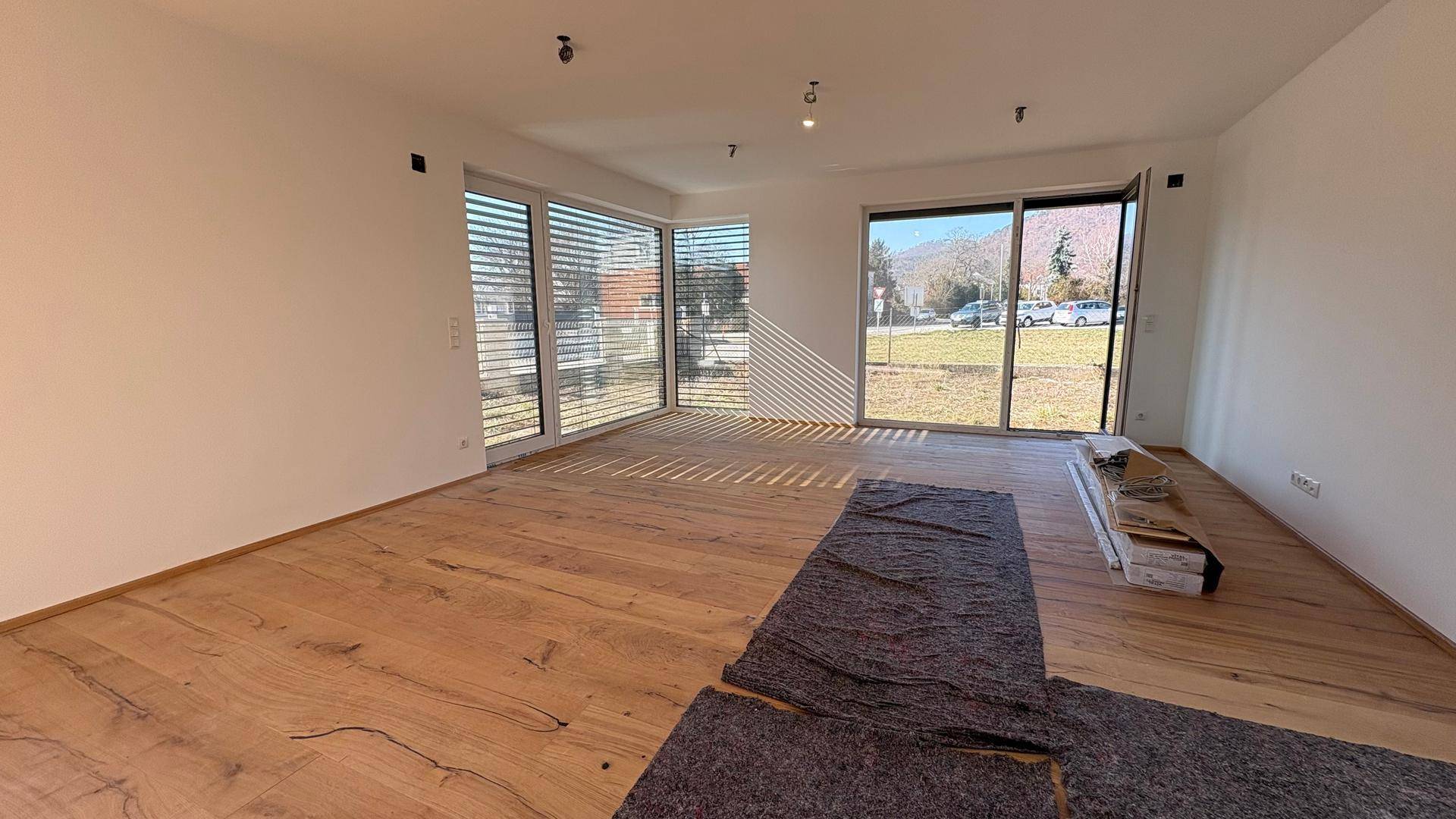 Heller, unmöblierter Wohnbereich mit großen Fenstern, Holzboden und Blick ins Freie.