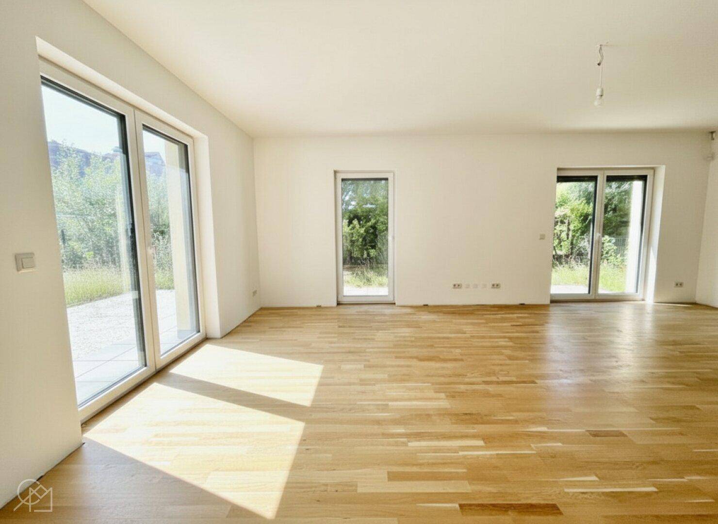 Heller, leerer Raum mit großen Fenstern, Holzboden und Zugang zum Garten.