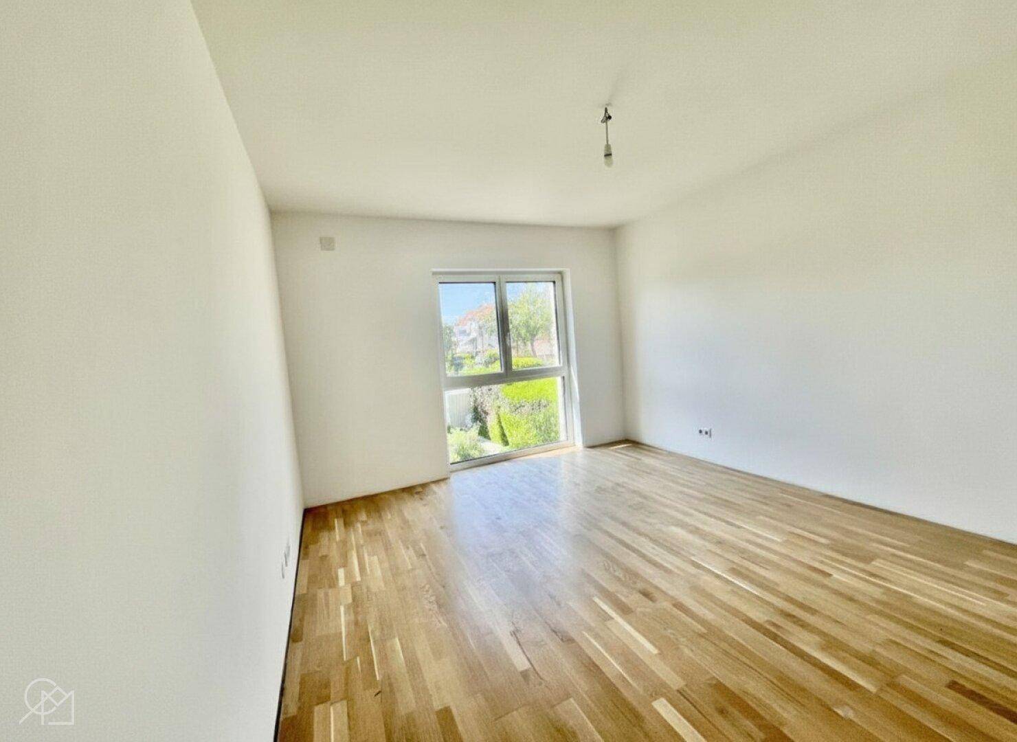 Helles, leeres Schlafzimmer mit Holzboden und großem Fenster mit Blick ins Grüne.
