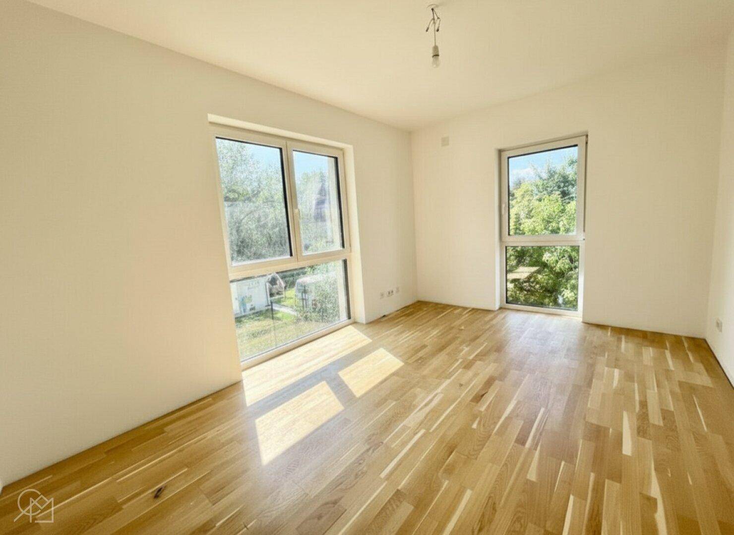 Heller, leerer Raum mit zwei großen Fenstern, Holzboden und viel Tageslicht.