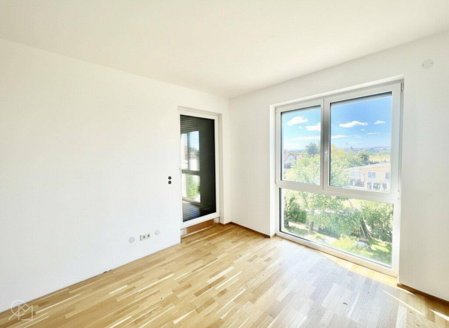 Heller, leerer Raum mit Holzboden, Fenster und Zugang zu einem Balkon.