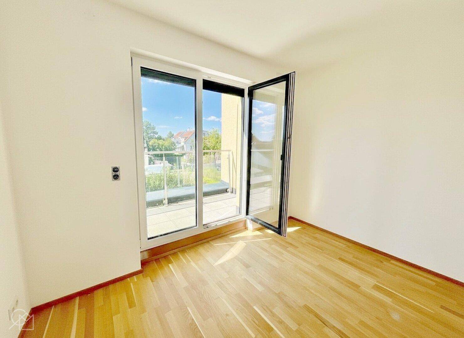 Heller, leerer Raum mit Holzboden und direktem Zugang zu einem sonnigen Balkon.
