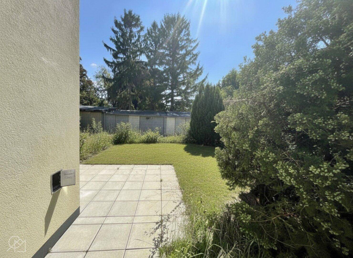 Gepflegter Gartenbereich mit Rasenfläche und gepflasterter Terrasse unter blauem Himmel.