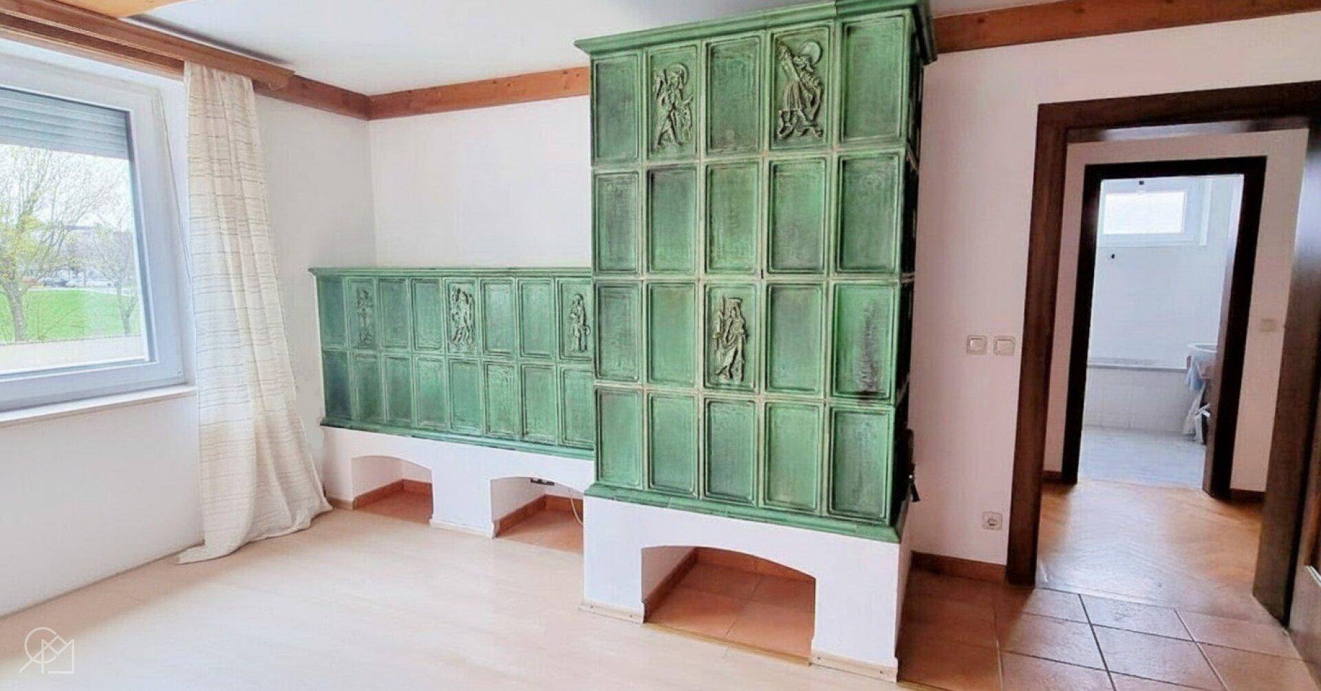 Unmöbliertes Zimmer mit großem grünem Kachelofen und Holzbalken.