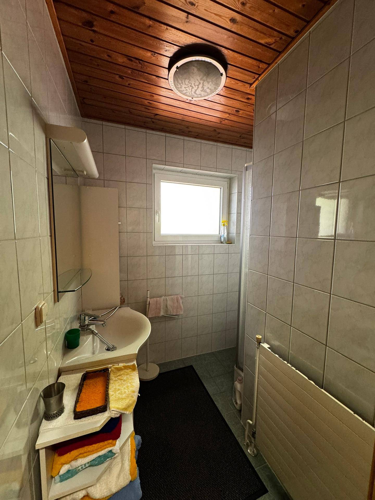 Badezimmer mit hellgrauen Fliesen, Dusche, Waschbecken und einem Fenster für natürliche Belüftung.