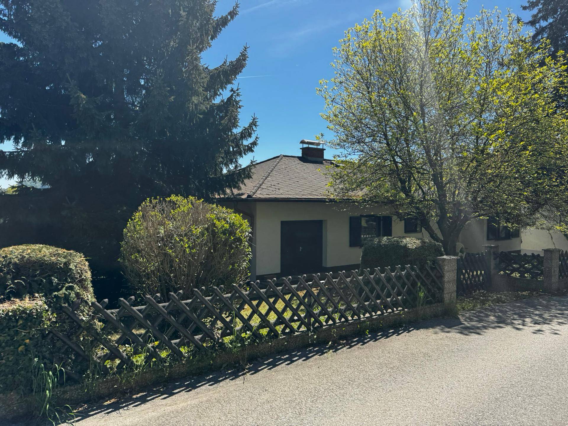 Ansicht des Hauses von der Straße aus, umgeben von Bäumen und einem Holzzaun.