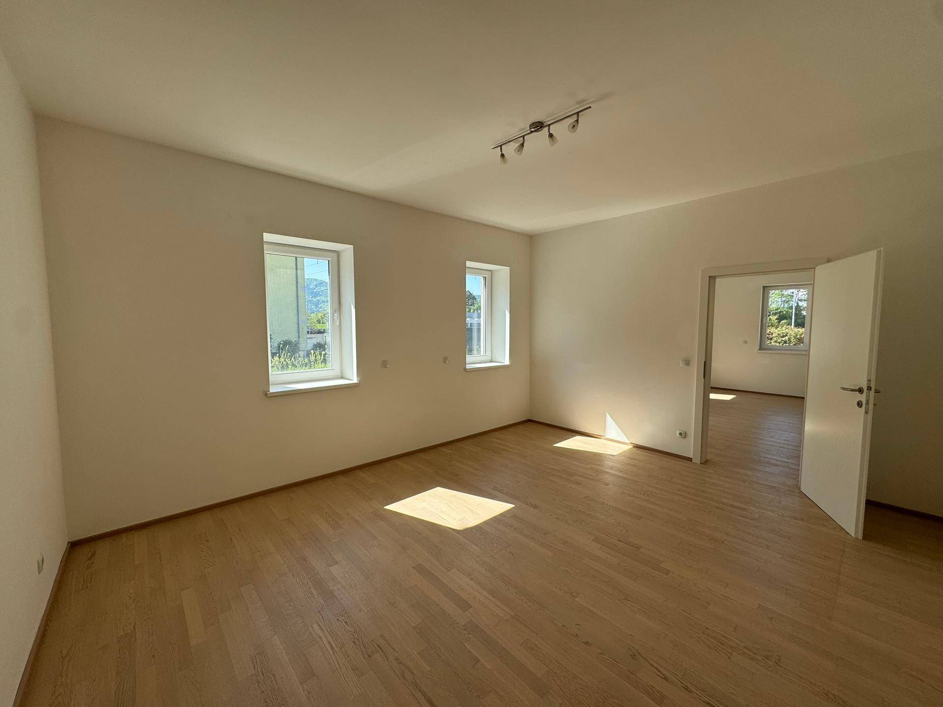 Geräumiges Wohnzimmer mit hellem Holzboden, weißen Wänden und viel natürlichem Licht durch die Fenster.