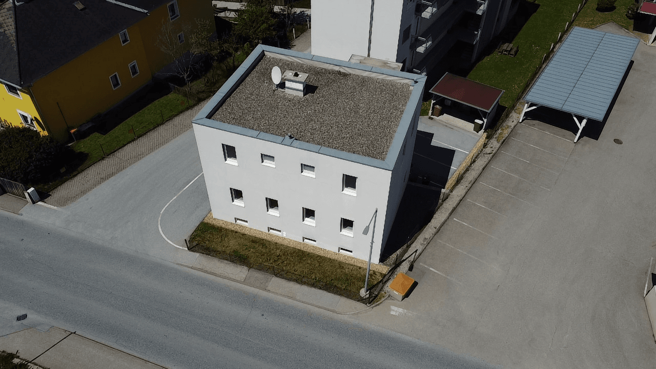 Luftaufnahme eines modernen Gebäudes mit Flachdach, umgeben von Parkplätzen und Grünflächen.