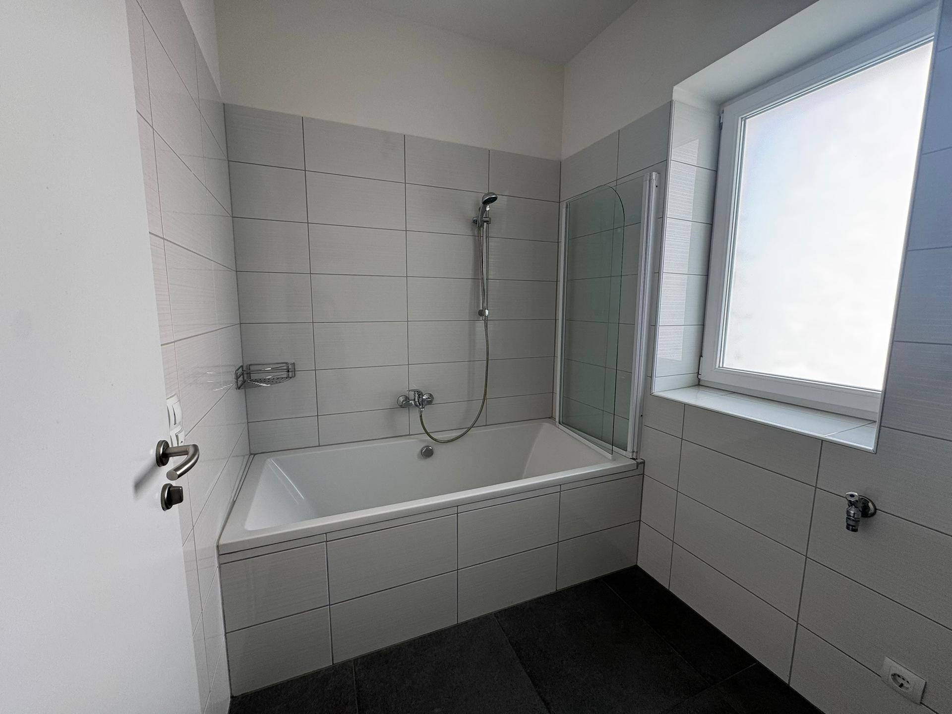 Helles Badezimmer mit Badewanne, weißen Fliesen und Fenster für natürliche Belüftung.