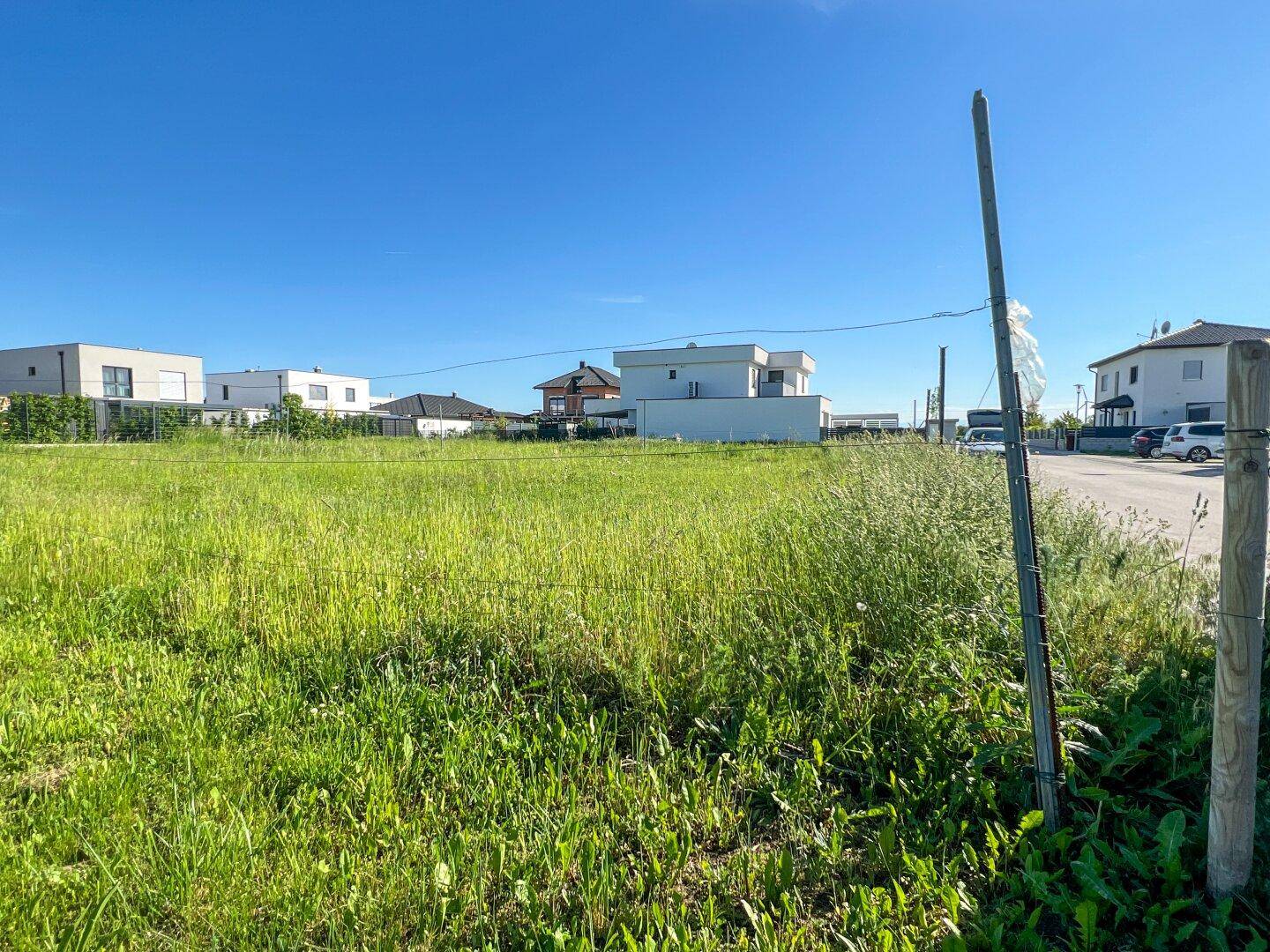 Offenes, grünes Grundstück mit angrenzender Straße und modernen Gebäuden im Hintergrund.