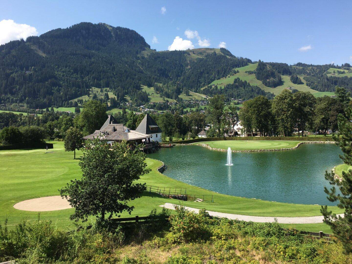 Idyllischer Golfplatz mit See, Springbrunnen und Bergblick unter blauem Himmel.