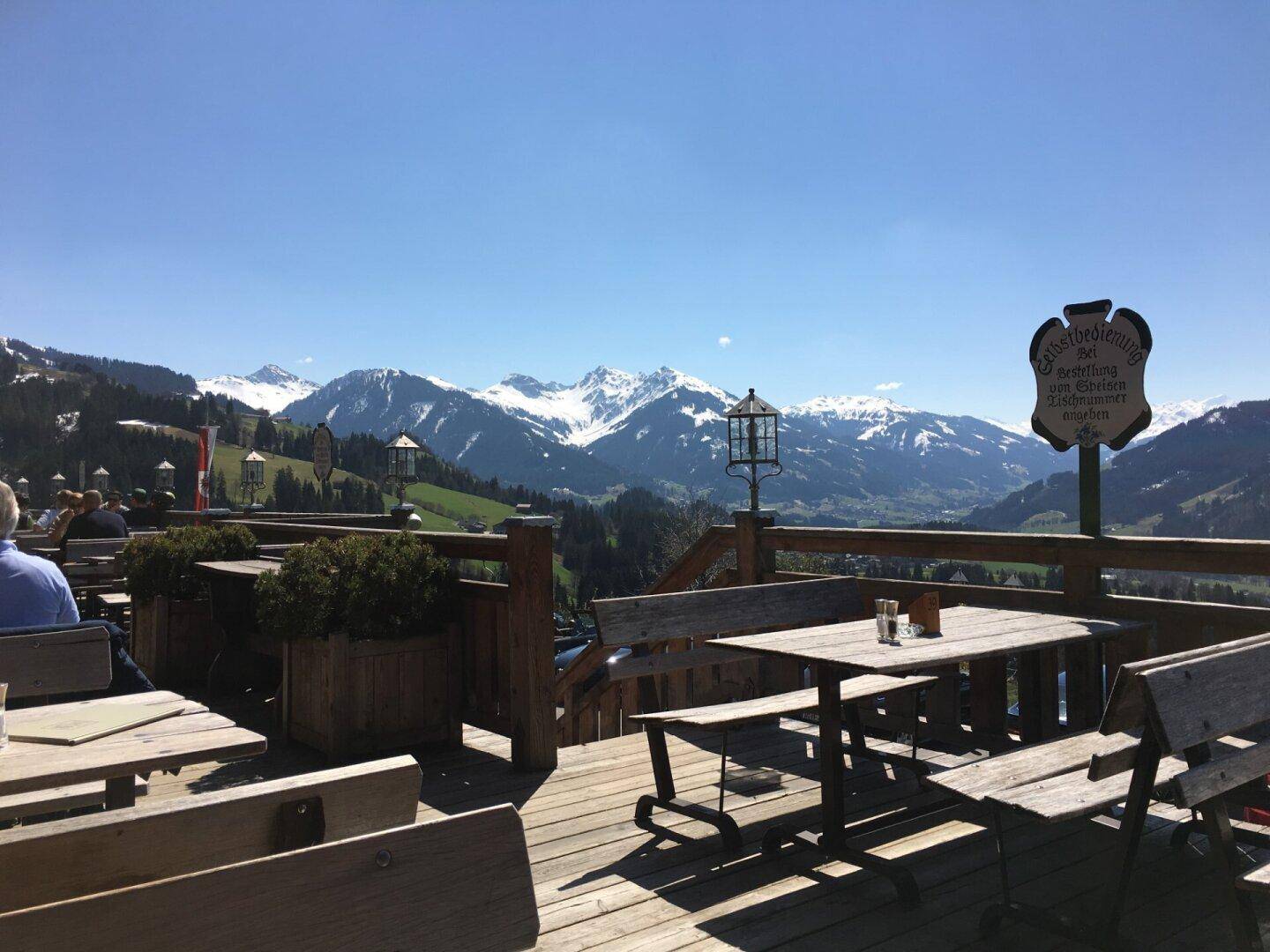 Sonnige Terrasse mit Holzbänken und Tischen, umgeben von einer beeindruckenden Bergkulisse.