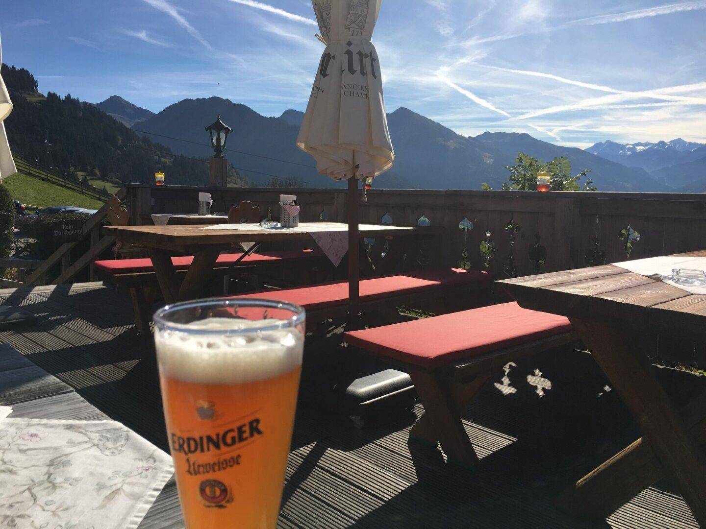 Sonnige Terrasse mit Holztischen und Bänken, einem Bierglas und Bergblick.