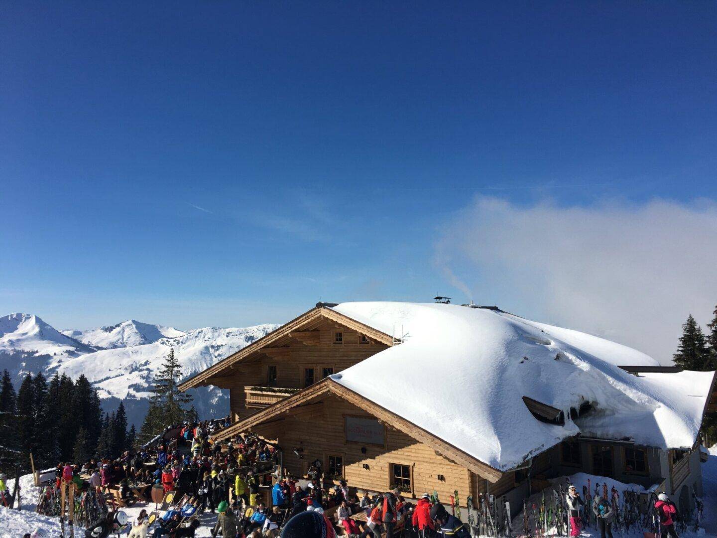 Belebte Skihütte mit viel Schnee und Menschen vor einer beeindruckenden Bergkulisse.