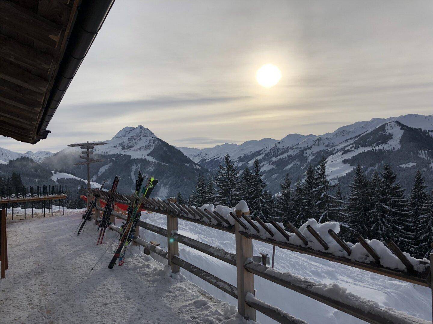 Winterlicher Ausblick von einer Terrasse auf verschneite Berge und Wälder bei Sonnenuntergang.